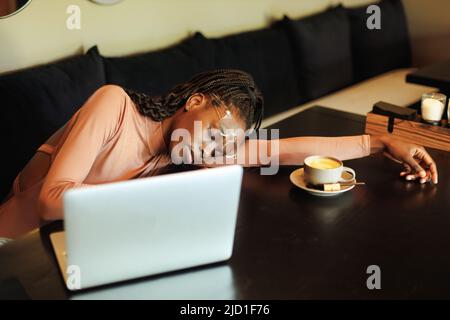 Müde multikulturelle afroamerikanische Frau in Brille, liegend und schluchzend auf dem Tisch am Laptop im Internetcafe mit Kaffee Stockfoto