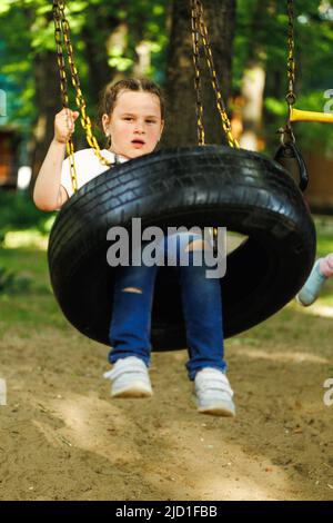 Coole kleine Mädchen schwingt auf Auto Gummirad mit gekreuzten Armen auf Spielplatz im grünen Park. Sommerferien im Camp, Touristenzentrum. Spielen im Freien Stockfoto