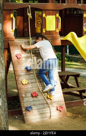 Kleines Mädchen klettert Seil und Step up Sportspiel-Komplex auf dem Spielplatz. Sommerferien im Camp, Touristenzentrum. Wandern und Spielen im Freien, Sport Stockfoto