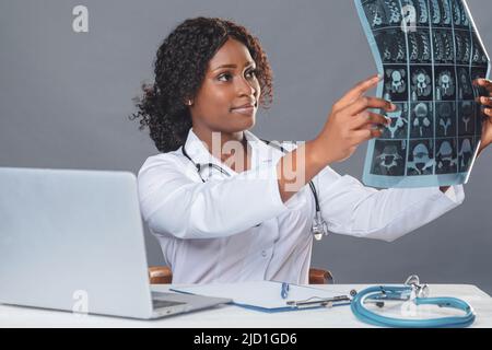 Afrikanische Ärztin in einem weißen Mantel und sitzt an einem Tisch in einem medizinischen Büro und arbeitet an einem Computer, Telemedizin, Online-Patientenberatung Stockfoto