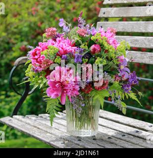 Buntes Bouquet in roten, rosa und violetten Farbtönen mit Pfingstrosen und Säulchen, steht in Glasvase auf dekorativer Holzgartenbank Stockfoto