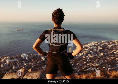 Rückansicht einer Wandererin, die auf dem Gipfel des Berges steht und auf das Stadtbild blickt Stockfoto