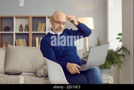 Seriöser älterer Mann, der zu Hause auf einem Stuhl sitzt, seinen Laptop benutzt und nachdenkt Stockfoto