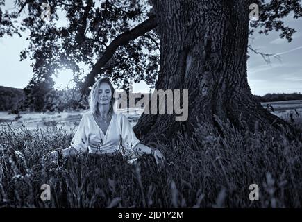 Monochromes Porträt einer jungen Frau, die bei Sonnenuntergang unter einer hundertjährigen Eiche Yoga-Meditation praktiziert Stockfoto