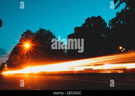 Die Scheinwerfer des Autos Streifen von Licht auf einer belebten Straße mit Straßenlaterne, Bäumen und blauem Himmel in der Nacht. Stockfoto