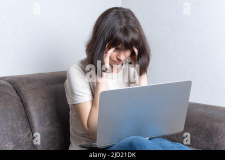 Frustrierte, traurige Frau mittleren Alters fühlt sich müde, massiert ihre Schläfen, sorgt sich um ein Problem, während sie mit einem Laptop auf einem Sofa sitzt. Arbeitsüberstunden Stockfoto