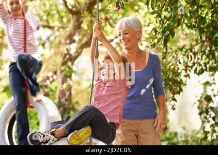 So machen Sie es. Aufnahme einer Frau, die ihrem Enkel auf einem Reifenschaukel mit ihrer Enkelin im Hintergrund hilft. Stockfoto