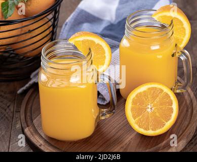 Zwei Gläser Orangensaft mit orangen Scheiben auf einer Holzplatte mit einem Korb mit Orangen im Hintergrund Stockfoto