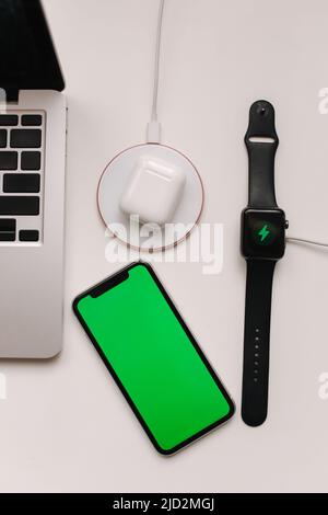 Arbeitsplatz mit Laptop auf Tisch und Geräte. Kabelloses Aufladen von Smart Watch und kabellosen Kopfhörern auf weißem Tisch. Smartphone mit grünem Bildschirm Stockfoto