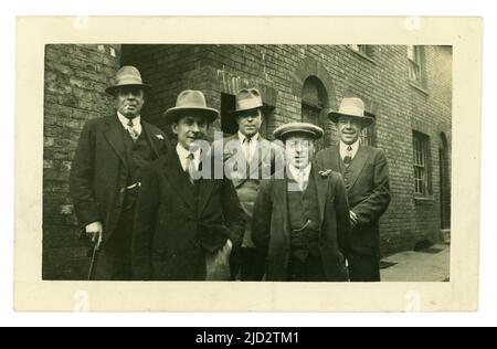 Ursprüngliche Postkarte aus den frühen 1920er Jahren einer Gruppe von Männern der Arbeiterklasse, die für ein Foto in einer städtischen Straße, in einer armen Nachbarschaft posieren, Fedora- oder Trilby-Hüte tragen und ein Mann eine flache Mütze, Peaky-Blinders/Gangster-Lookalikes, möglicherweise eine Hochzeitsfeier trägt, Bleibt zurück, während der Mann vorne eine Knopflochblume auf dem Revers trägt. Ca. 1925. GROSSBRITANNIEN Stockfoto