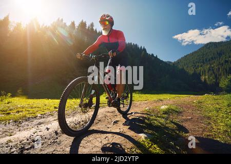 Der alte Radler mit weißem Bart fährt mit seinem Mountainbike im grünen Fichtenwald in rotem Kostüm in Almaty, Kasachstan. Extremsport und Outdoor-Erholung Stockfoto