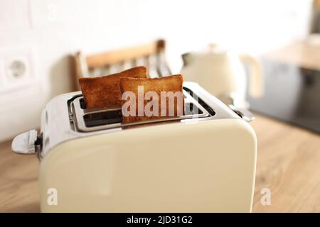 Geröstetes Brot und Toaster zum Frühstück. Moderner weißer Toaster und geröstete Brotscheiben im Inneren auf einem Holztisch in der Küche Stockfoto
