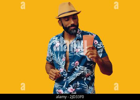 Glücklicher afroamerikanischer Mann mit mittlerem Erwachsenen, der ein hawaiianisches Hemd mit Cocktailglas und einem Hut im Studio trägt Stockfoto