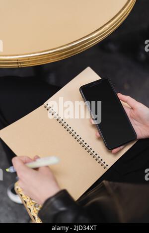 Aus dem hohen Winkel betrachtet, wie eine Frau das Smartphone mit leerem Bildschirm und ein Notebook in der Nähe des runden Tisches hält Stockfoto