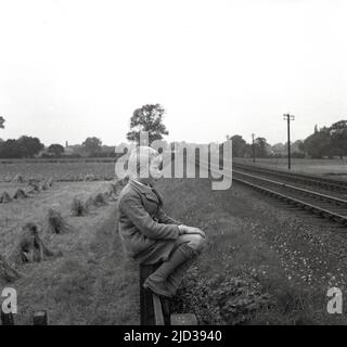 1950s, historisch, ein junger Schuljunge in seiner Jacke und Shorts, auf einem Holzzaun neben einem Feld neben einer Eisenbahnlinie sitzend, wartet auf einen Dampfzug, der mitkommt, England, Großbritannien. Stockfoto