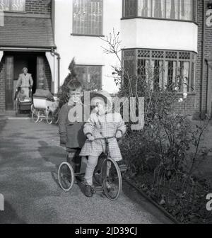 1950s, historisch, auf einem Eingangsweg zu einem Haus im Vorort Bramhall, Stockport, England, Großbritannien, eine Mutter mit einem Kinderwagen und Kinderwagen an der Eingangstür, mit ihren zwei kleinen Kindern, eines davon auf einem Metalldreirad. Stockfoto