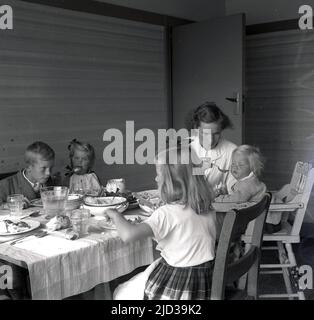 1940s, historisch, eine Dame mit ihren kleinen Kindern, die an einem Tisch essen, Bramhall, Stockport, England, Großbritannien. Das jüngste Kind, ein Kleinkind, sitzt auf einem hölzernen Hochstuhl und wird von ihrer Mutter gefüttert. Stockfoto