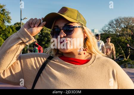 Porträt einer hübschen jungen lateinamerikanischen Argentinierin, plus-size-Modell, im Freien bei Sonnenuntergang in der Stadt, stehend mit Brille, Mütze und Blick auf den Kameradinnen Stockfoto