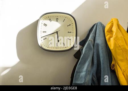 Eine Uhr im Schatten und Sonnenlicht an der Wand eines Flurs mit Mänteln an Kleiderhaken Stockfoto