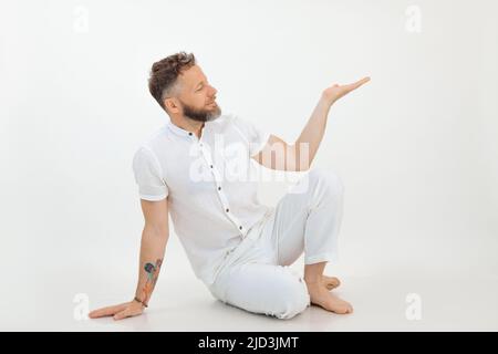 Lächelnder Geschäftsmann mit Tattoo sitzt auf dem Boden in weißem Outfit auf weißem Hintergrund. Werbemanager, Präsentation Stockfoto