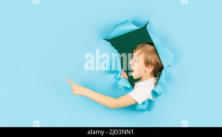 Junge in blauem Papierloch, der mit dem Finger zur Seite zeigt. Schreit Junge, der durch ein Loch in der Papierwand schaut. Schockiert Kind macht Loch in Papier. Kinderlooks Stockfoto