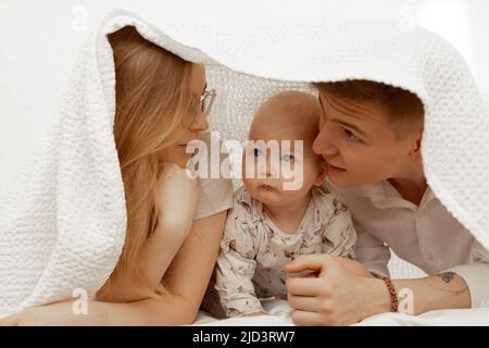 Junge Mutter und Vater lagen unter einer weißen Decke mit einem kleinen Baby auf dem Bett und genießen. Nette Beziehung zwischen Eltern und Kleinkind Nahaufnahme, kostenlos Stockfoto