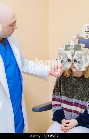 Ein männlicher Augenarzt überprüft das Augenlicht eines Mädchens mit einem Phoropter. Sehbehandlung. Stockfoto