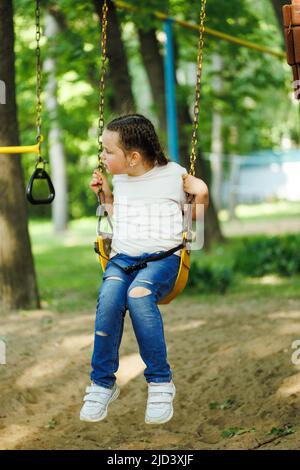 Nettes kleines Mädchen, das auf gelber Schaukel auf dem Spielplatz im grünen Park wimmelt. Sommerferien im Camp, Touristenzentrum. Wandern und Spielen im Freien, Sport Stockfoto