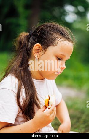 Seitenansicht eines kleinen, nachdenklichen Mädchens mit dunklen Haaren, das Sandwich hält, kaut und im Parkwald zwischen grünen Bäumen sitzt. Stockfoto