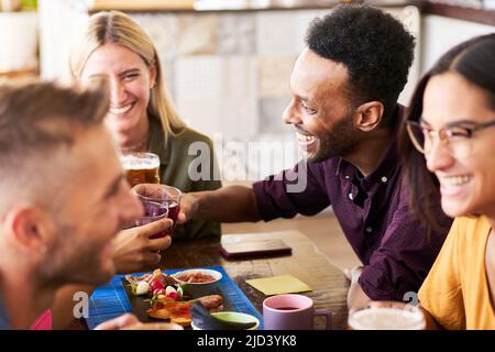 Eine Gruppe von multikulturellen Leuten trinkt Bier in einer Bar. 30s Junge Freunde, die Spaß haben und in einer Happy Hour auf dem Dach zu Mittag essen und Alkohol trinken. Stockfoto