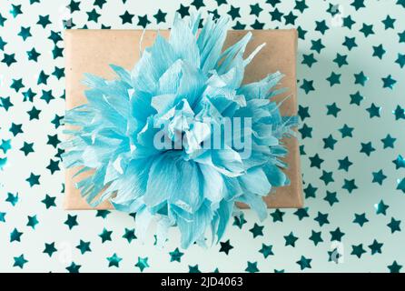 Geschenkbox aus Kraftpapier mit blauer Schleife auf blauem Hintergrund mit Konfetti. Weihnachtsgeschenk, valentinsüberraschung, Geburtstagskonzept. Flach liegend Stockfoto