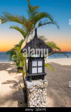 Schwarze Laterne aus Holz mit schwarzer Strohspitze und weißen Reisfeldern im japanischen Stil, die auf einem Kopfsteinpflaster auf dem Sand mit Palmen und O sitzen Stockfoto