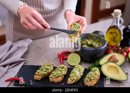 Frau, die Bruschetta mit frisch zubereiteter Guacamole in der heimischen Küche macht Stockfoto