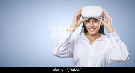 Virtual-Reality-Konzept: Lächelte junge Frau in hellem Hemd mit VR-Brille auf dem Kopf auf hellblauem Hintergrund mit Platz für Ihr Logo oder Text, verspottet Stockfoto