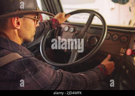 Kaukasischer Cowboy-LKW-Fahrer mit westlicher Mütze und Sonnenbrille, der in seinem alten Semi-Truck auf die Straße geht und bereit für eine lange Reise ist. Stockfoto