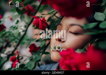 Porträt einer jungen asiatischen Frau mit geschlossenen Augen und Kamillenblüten im Mund, die sich bei blühenden roten Kletterrosen im Garten vergnügt. Speicherplatz kopieren. Stockfoto