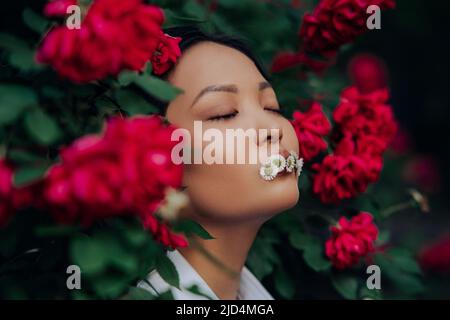 Porträt einer jungen asiatischen Frau mit geschlossenen Augen und Kamillenblüten im Mund, die sich bei blühenden roten Kletterrosen im Garten vergnügt. Stockfoto