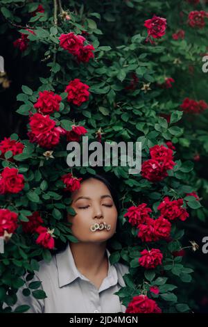 Porträt einer jungen asiatischen Frau mit geschlossenen Augen und Kamillenblüten im Mund, die sich bei blühenden roten Kletterrosen im Garten vergnügt. Stockfoto