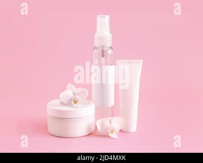 Weißes Kosmetikglas, Tube und Flasche in der Nähe von weißer Orchideenblume auf hellrosa, Nahaufnahme, Mockup. Hautpflegeprodukt, Creme oder Lotion. Exotisch natürlich Stockfoto