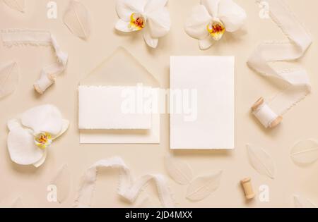 Hochzeitskarten in der Nähe von weißen Orchideenblumen und Seidenbändern auf hellgelber Draufsicht, Mockup. Romantische Szene mit leerer Karte flach legen, Platz für Text. Stockfoto