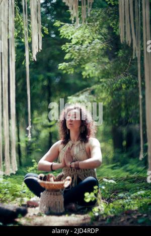 Eine Frau mit dunklen lockigen Haaren, die als Einheimische gekleidet ist, meditiert im Wald Stockfoto