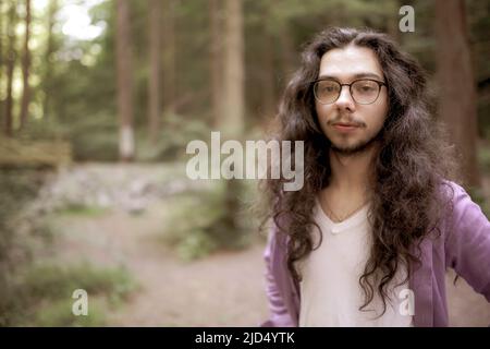 Ein hübscher junger Mann mit langen Haaren steht abseits der Mitte im Wald und blickt direkt auf die Kamera. Unfokussierte Hintergrundkopie. Stockfoto