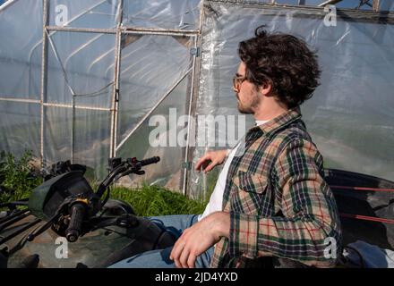 Junger kaukasusmännlicher Bauer auf einem Geländewagen, der neben einem Bio-Gemüsegarten-Gewächshaus in natürlicher goldener Stunde Sonnenlicht wi kontempliert Stockfoto
