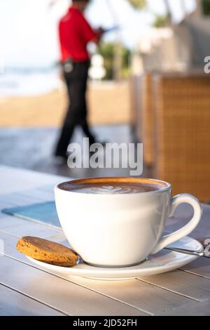 Nahaufnahme von Kaffee-Latte in einer weißen Tasse und Untertasse mit kleinem Shortbread-Cookie auf dem Tisch, während der Restaurantbearbeiter vor verschwommenem Hintergrund läuft Stockfoto