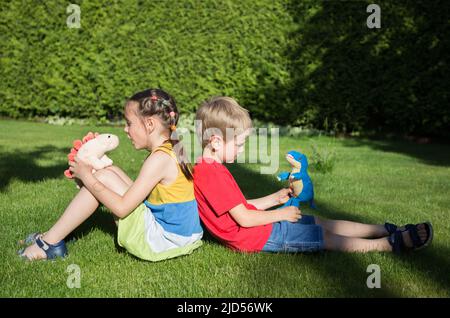 Junge und ein Mädchen von 6-7 Jahren sitzen zurück auf dem Gras im Garten. Spielen Sie Fantasy-Spiele mit Plüsch-Dinosaurier. Freundschaft, Kindheit, s Stockfoto