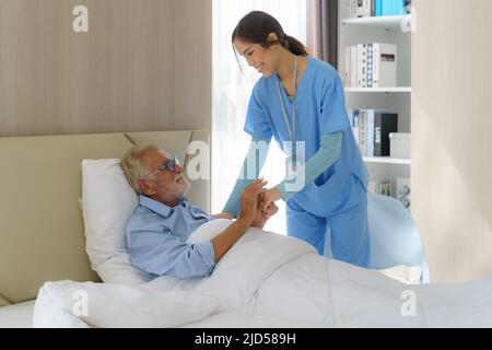 Asiatische Krankenschwester, die auf einem Heimbett neben einem älteren Mann steht, der den Händen und der Pflege hilft. Altenpflege und Gesundheit Lebensstil, medizinisches Konzept. Stockfoto