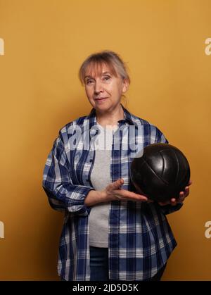 Lächelnde ältere Frau in einer legeren Form, die den Fußball vor gelbem Hintergrund hält Stockfoto