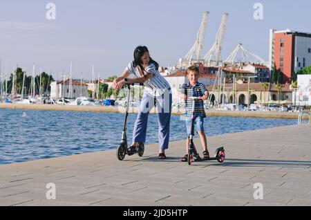 Fröhlicher Familienleben und Urlaubskonzept. Mutter, kleiner Junge, Mädchen, Roller reiten, in der Altstadt spazieren gehen, Straße. Lachen an einem sonnigen Sommertag. Stockfoto