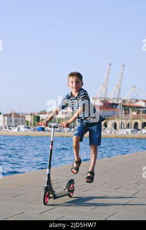 Urlaubskonzept. Kleiner Junge, der Roller reitet, in der Altstadt, Straße zu Fuß. Lachen an einem sonnigen Sommertag. Spaß haben Stockfoto