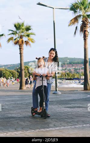 Fröhlicher Familienleben und Urlaubskonzept. Mutter, kleiner Junge, Mädchen, Roller reiten, in der Altstadt spazieren gehen, Straße. Lachen an einem sonnigen Sommertag. Stockfoto